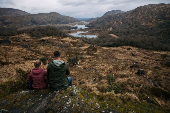 Een man en een vrouw zitten op een met mos begroeide rots, met hun rug naar de camera, uitkijkend over een uitgestrekte vallei met kronkelende rivieren, bruin gras, dichte bomen en bergen in de verte onder een bewolkte hemel. Het landschap oogt ruig, natuurlijk en sereen. | Echt Ierland Vakanties
