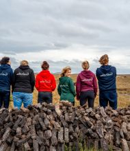 Vijf mensen in hoodies staan met hun rug naar de camera, kijkend naar een uitgestrekt, bewolkt landschap op hun vakantie in Ierland. Ze staan achter een grote stapel turfstenen. De hoodies, in blauw, rood, groen en zwart, gaan op in de buitenlucht terwijl de open velden zich voor hen uitstrekken. | Echt Ierland Vakanties