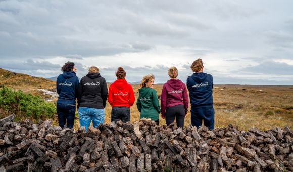 Vijf mensen in hoodies staan met hun rug naar de camera, kijkend naar een uitgestrekt, bewolkt landschap op hun vakantie in Ierland. Ze staan achter een grote stapel turfstenen. De hoodies, in blauw, rood, groen en zwart, gaan op in de buitenlucht terwijl de open velden zich voor hen uitstrekken. | Echt Ierland Vakanties