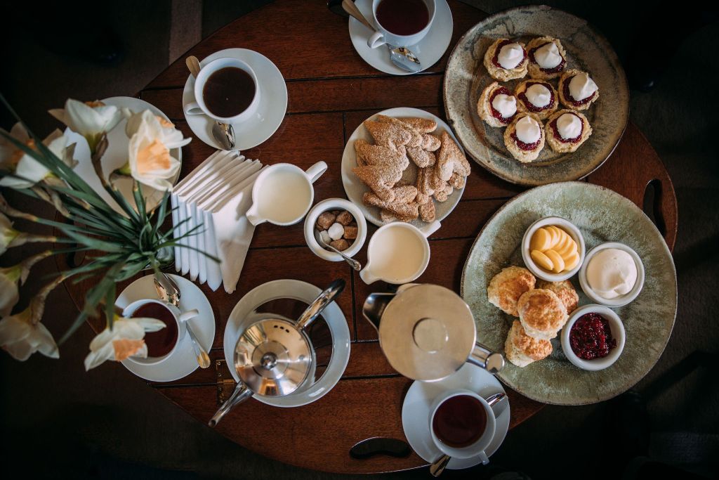 Een houten tafel is gedekt voor thee met kopjes zwarte thee, een theepot, melkkannetje en suikerklontjes. Een bord met scones met jam en room, gebak in de vorm van schelpen en een schaal met boter zijn ook aanwezig. Een vaas met witte bloemen en gevouwen servetten voegen een decoratief tintje toe. | Echt Ierland Vakanties