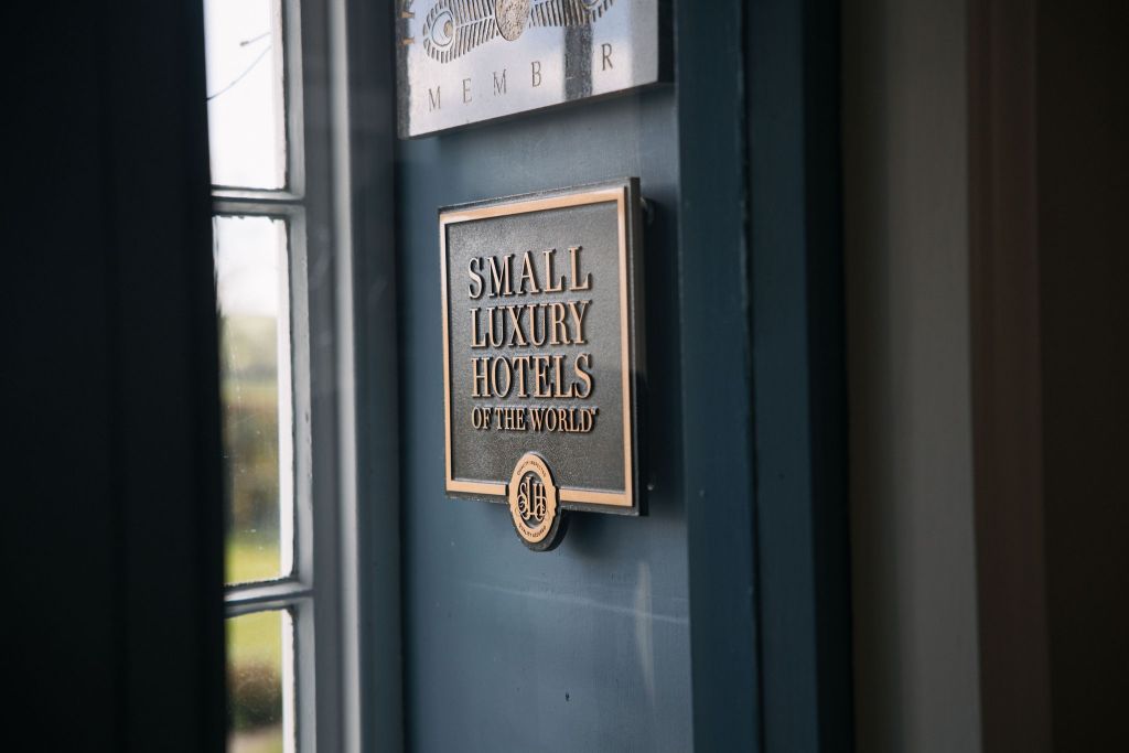 Plaquette op een blauwe deur met de tekst "Small Luxury Hotels of the World" met daaronder een embleem. Zonlicht filtert door een raam aan de linkerkant en werpt een zachte gloed op het metalen oppervlak van de plaquette. De scène suggereert een verfijnd en exclusief etablissement. | Echt Ierland Vakanties