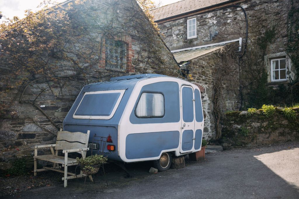 Een kleine, vintage blauw-witte camper staat geparkeerd naast een rustiek stenen huisje met overwoekerde klimop. Er staat een houten bankje bij de camper. Het tafereel baadt in zacht zonlicht, werpt zachte schaduwen en creëert een serene, nostalgische sfeer. | Echt Ierland Vakanties