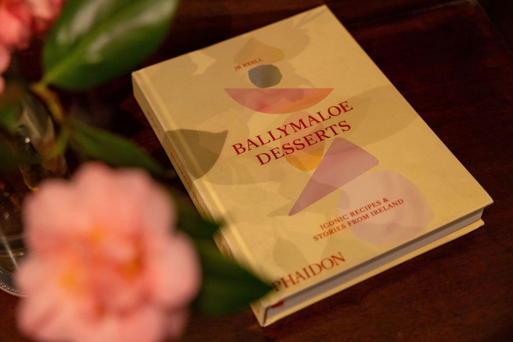 Een boek met de titel "Ballymaloe Desserts: Iconic Recipes & Stories from Ireland" rust op een houten oppervlak. De cover toont abstracte geometrische vormen in zachte kleuren. Op de voorgrond is links gedeeltelijk een vaas met roze bloemen en groene bladeren zichtbaar. | Echt Ierland Vakanties