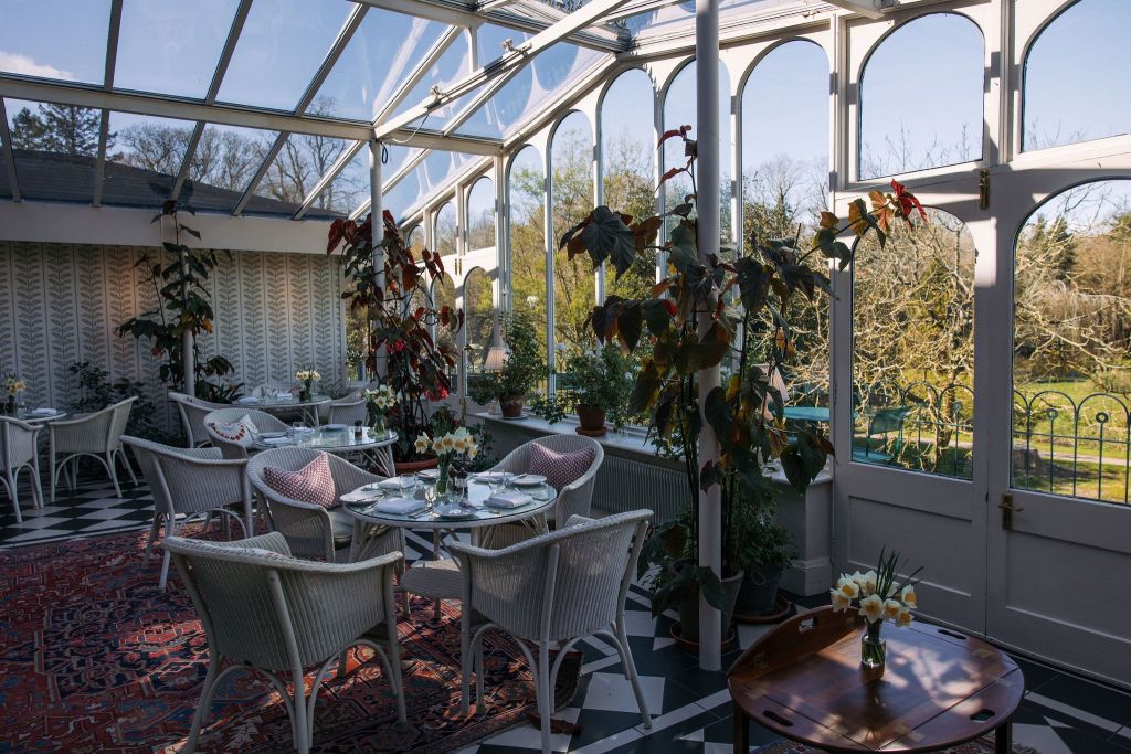Een zonnige serre met een glazen plafond en muren, met witte rieten stoelen rond ronde glazen tafels. Potplanten sieren de ruimte en een sierlijk tapijt bedekt een deel van de zwart-wit geblokte vloer. Door het glas is een tuin met bomen buiten zichtbaar. | Echt Ierland Vakanties