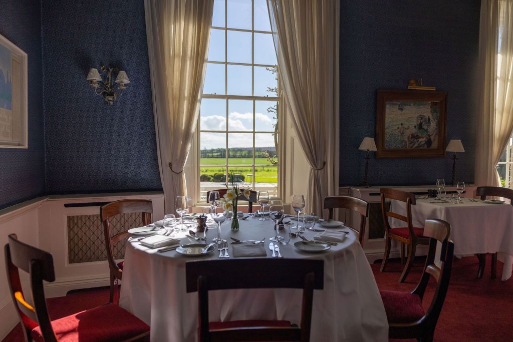 Elegante eetkamer met een ronde tafel voor zes personen, met wit tafelkleed, glaswerk en zilverwerk. Groot raam met doorschijnende gordijnen biedt uitzicht op een weelderig groen landschap onder een blauwe lucht. Muren zijn donker, versierd met schilderijen en de vloer is bedekt met rood tapijt. | Echt Ierland Vakanties