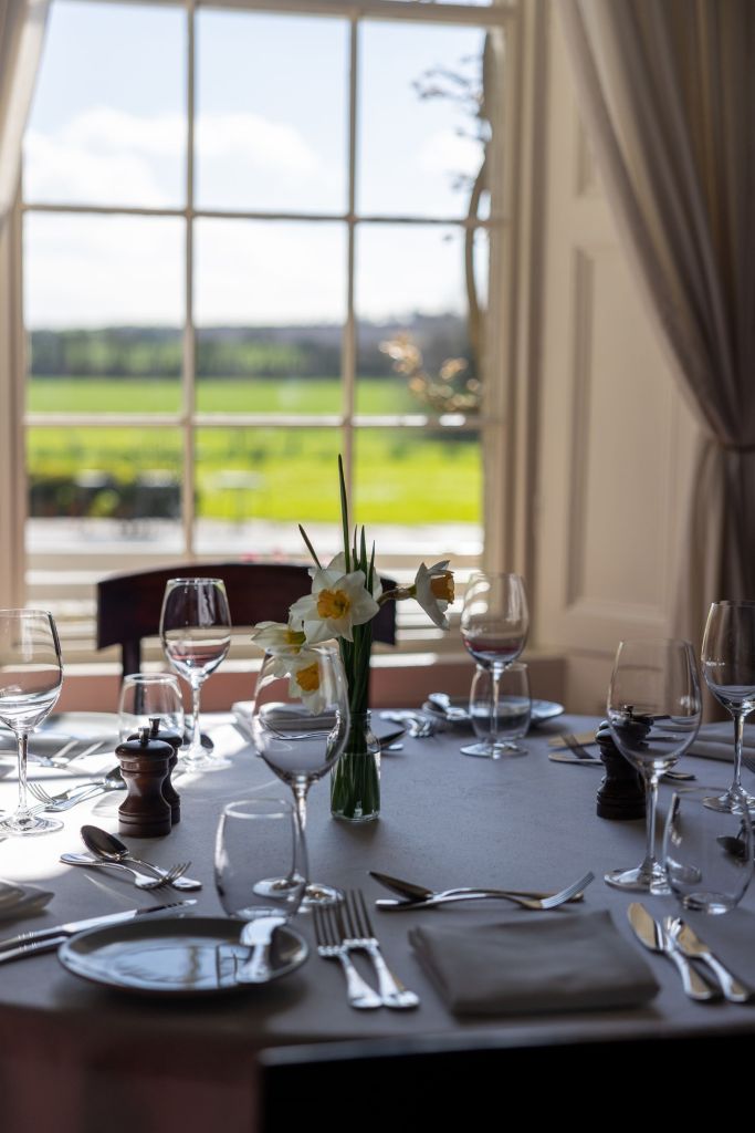 Elegante eettafelset met glaswerk, bestek en borden met servetten. Een vaas met narcissen dient als middelpunt. Zonlicht filtert door grote ramen met uitzicht op een groen buitenlandschap en een heldere lucht. Crèmekleurige gordijnen omlijsten het raam. | Echt Ierland Vakanties