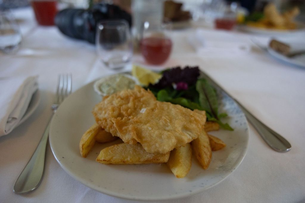 Een close-up van een gerecht met gepaneerde vis op dikke frieten, vergezeld door een klein beetje tartaarsaus. Een partje citroen en gemengde groenten, waaronder sla en een plakje komkommer, maken het bord compleet. De tafel is gedekt met een vork, mes en servet op een wit tafelkleed. | Echt Ierland Vakanties