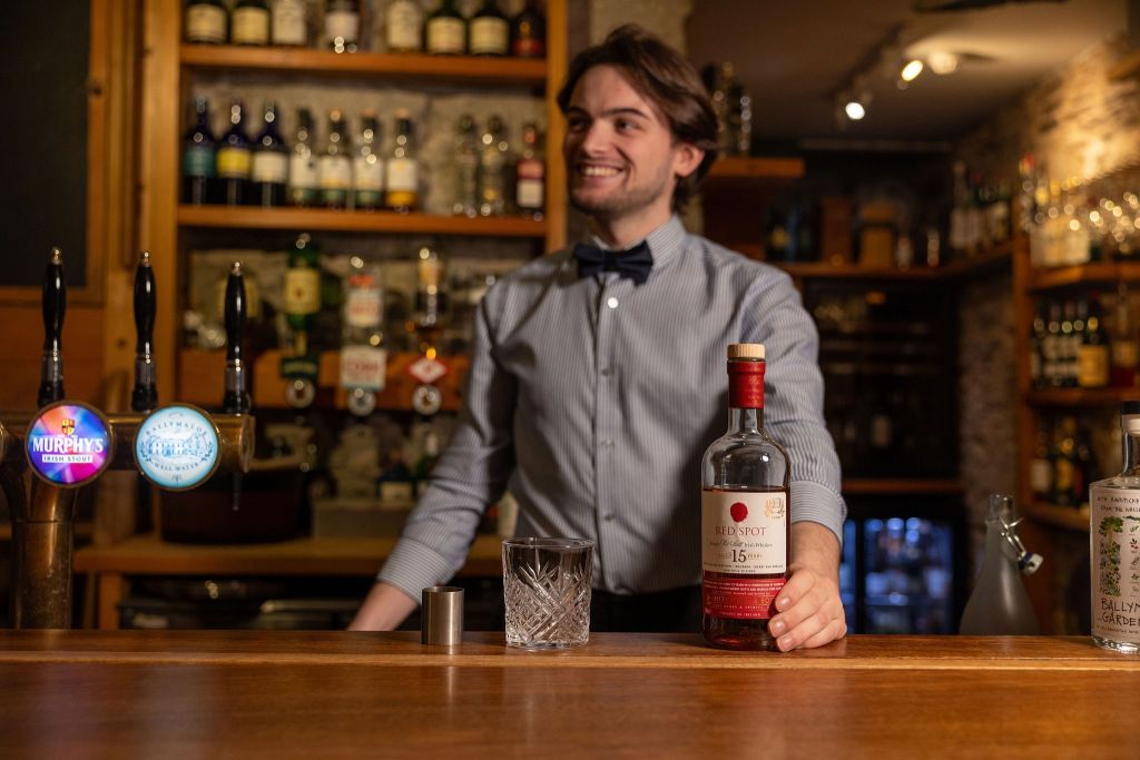 Een lachende barman in een gestreept overhemd en vlinderdas staat achter een houten bar. Hij houdt een fles Redbreast 15-year-old whisky vast naast een kristallen glas. De achtergrond van de bar bestaat uit verschillende flessen en twee biertaps, waarvan er één is gelabeld als Murphy's. Gezellige verlichting voegt warmte toe aan de setting. | Echt Ierland Vakanties