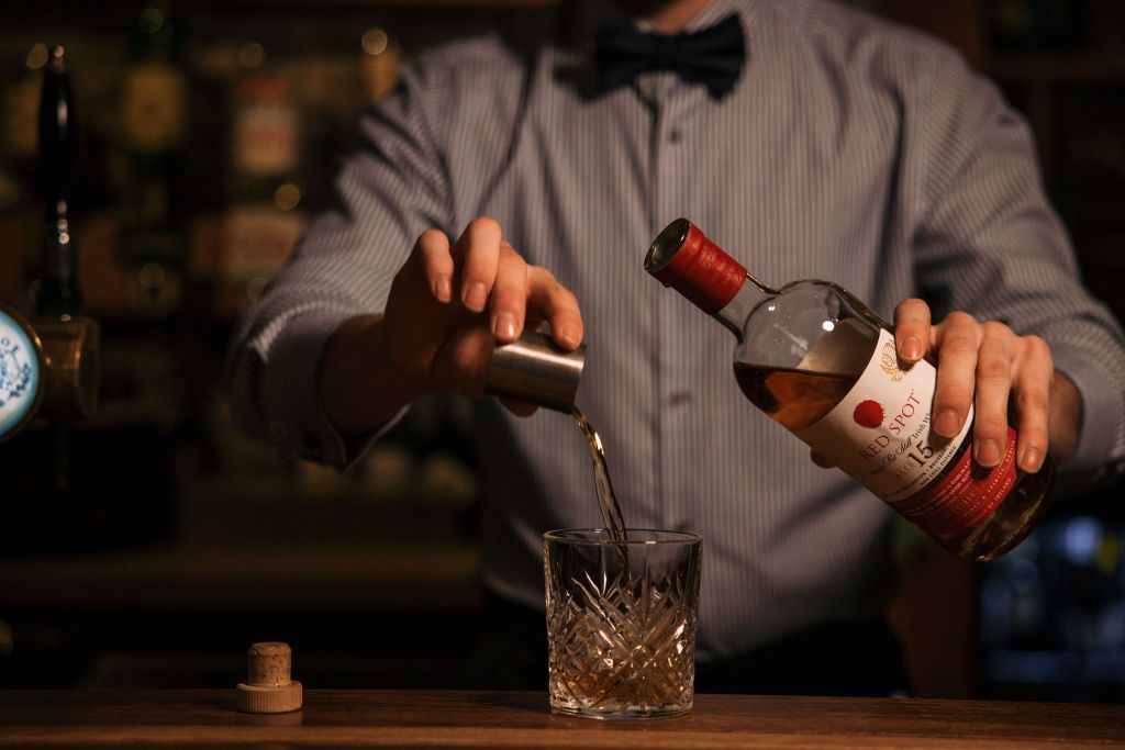 Een barman in een gestreept overhemd en vlinderdasje schenkt whisky uit een fles met het label "Red Spot" in een jigger, die in een geslepen glazen tumbler giet. De kurk rust op de houten bar. De achtergrond is zwak verlicht met wazige flessen. | Echt Ierland Vakanties