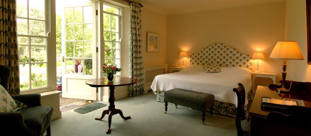 Een gezellige slaapkamer met een groot bed met een hoofdeinde met patroon. Twee lampen en een ingelijste foto hangen boven nachtkastjes aan beide kanten. Er staat een ronde tafel met een bloemenvaas in de buurt en een comfortabele stoel. Grote ramen met gordijnen met patroon geven uitzicht op een tuin met groen. | Echt Ierland Vakanties