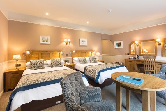 Gezellige hotelkamer met twee tweepersoonsbedden, elk versierd met kussens met patroon en een marineblauwe sprei. Warme verlichting van wandlampen en tafellampen verlicht de ruimte. Een houten bureau met een stoel en een ronde tafel met fauteuils voegen functionaliteit toe. Beige muren tonen ingelijste kunst. | Echt Ierland Vakanties