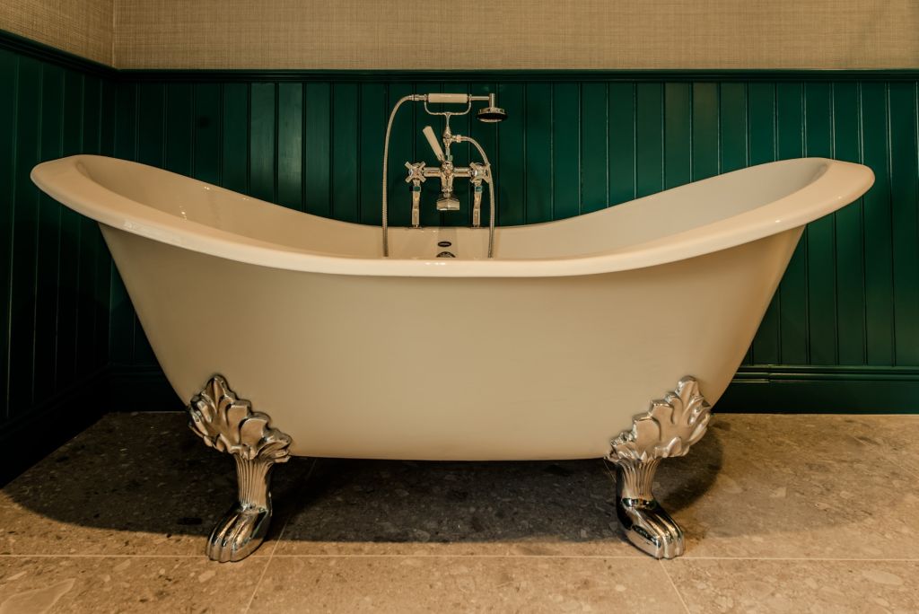 Een vrijstaand bad in vintage-stijl met glanzende pootjes op pootjes, met sierlijke ontwerpen, staat op een betegelde vloer. Het bad staat tegen een muur met donkergroene panelen en bevat gepolijste zilveren kranen en een handdouche. De setting is elegant en klassiek. | Echt Ierland Vakanties