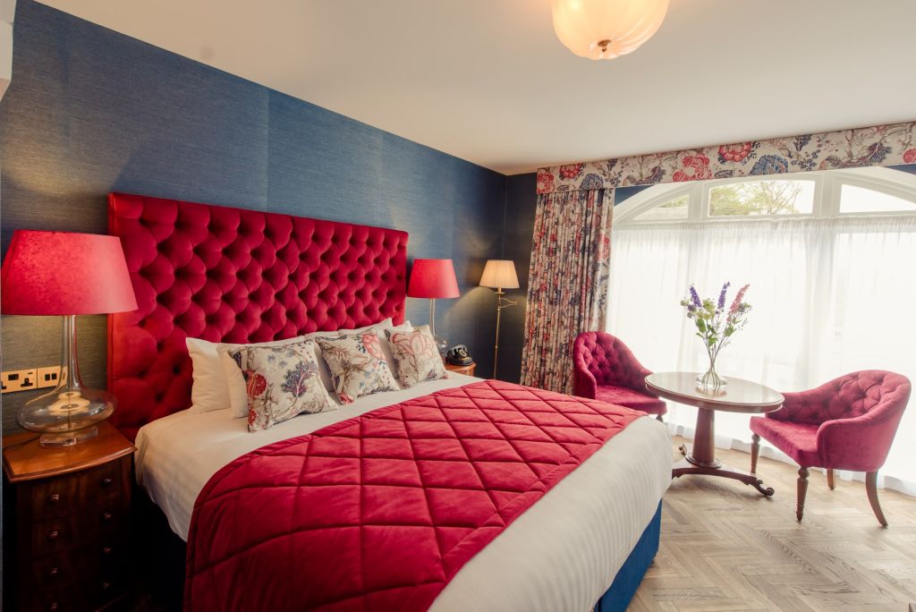 Elegante hotelkamer met een groot bed met een gecapitonneerd rood hoofdeinde, bijpassende rode quilt en kussens met bloemenaccenten. Twee rode lampen staan op nachtkastjes. Een ronde tafel met bloemen en twee rode stoelen staan bij een raam met bloemengordijnen. De muren zijn blauw en de vloer is van hout. | Echt Ierland Vakanties