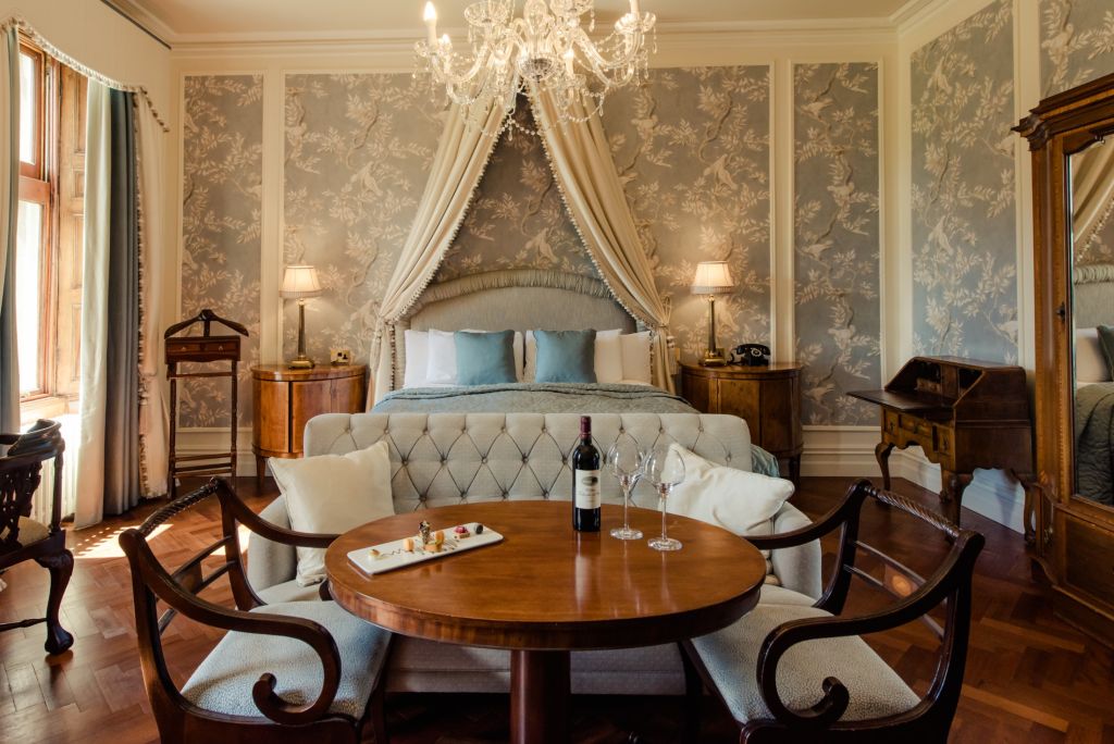 Elegante slaapkamer met een kroonluchter, behang met bloemenmotief en een hemelbed met grijs en blauwgroen beddengoed. Een kleine ronde tafel met een fles wijn, twee glazen en een dienblad met hapjes staat op de voorgrond. Vintage houten meubels en zachte verlichting creëren een gezellige sfeer. | Echt Ierland Vakanties