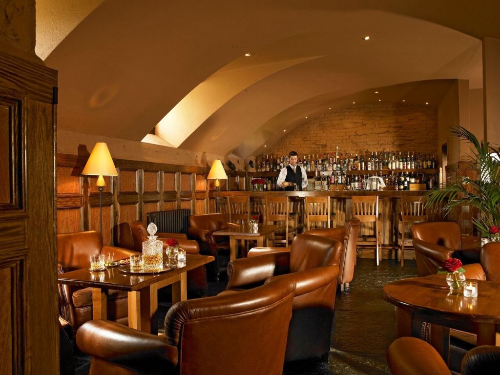 Gezellige lounge met gedimd licht, met leren stoelen en houten tafels. Een barman staat achter een goed gevulde bar met een stenen achtergrond. Zachte lampen voegen warmte toe. De setting is intiem, met netjes gerangschikte tafels en kleine decoratieve details zoals een roos en een glazen karaf. | Echt Ierland Vakanties
