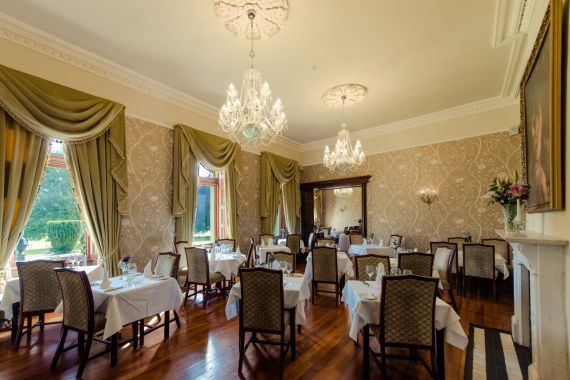 Elegante eetkamer met sierlijke kroonluchters, groene gordijnen en behang met patronen. Verschillende houten tafels met witte tafellakens en stoelen met kussens zijn netjes gerangschikt. Grote ramen bieden uitzicht op een weelderige tuin. Een spiegel en decoratieve lampen versterken de luxueuze ambiance. | Echt Ierland Vakanties