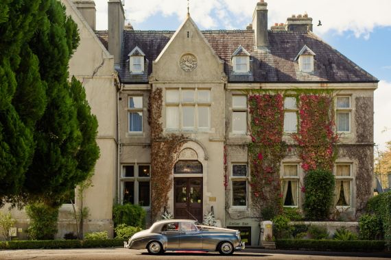 Een oldtimer staat geparkeerd voor een groot, historisch landhuis versierd met klimop. Het landhuis heeft meerdere schoorstenen, een stenen gevel en verschillende ramen met een symmetrisch ontwerp. Een grote boom omlijst gedeeltelijk de linkerkant, terwijl de lucht grotendeels helder is met een paar wolken. | Echt Ierland Vakanties