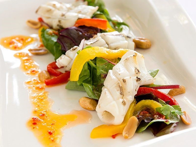 Een kleurrijk gerecht met gegrilde inktvisringen met verkoolde plekken, op een bedje van gemengde groenten, rode en gele paprikareepjes en amandelen. Een levendige oranje saus met zichtbare chilivlokken is op artistieke wijze over het witte rechthoekige bord gedruppeld. | Echt Ierland Vakanties