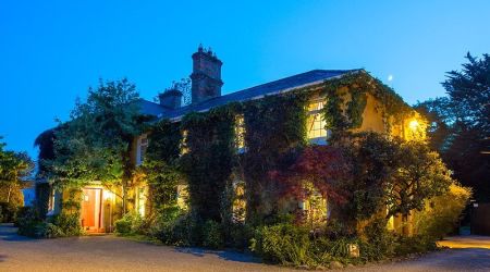 Verlicht groot huis in de schemering, omgeven door klimop en weelderig groen. Warm licht van ramen en open deur creëert een gastvrije sfeer. Schoorstenen zichtbaar op het dak. De lucht is helder en blauw, wat de gezellige ambiance versterkt. Grind oprit en goed onderhouden tuin aan de voorkant. | Echt Ierland Vakanties