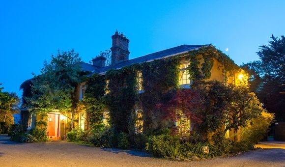 Verlicht groot huis in de schemering, omgeven door klimop en weelderig groen. Warm licht van ramen en open deur creëert een gastvrije sfeer. Schoorstenen zichtbaar op het dak. De lucht is helder en blauw, wat de gezellige ambiance versterkt. Grind oprit en goed onderhouden tuin aan de voorkant. | Echt Ierland Vakanties