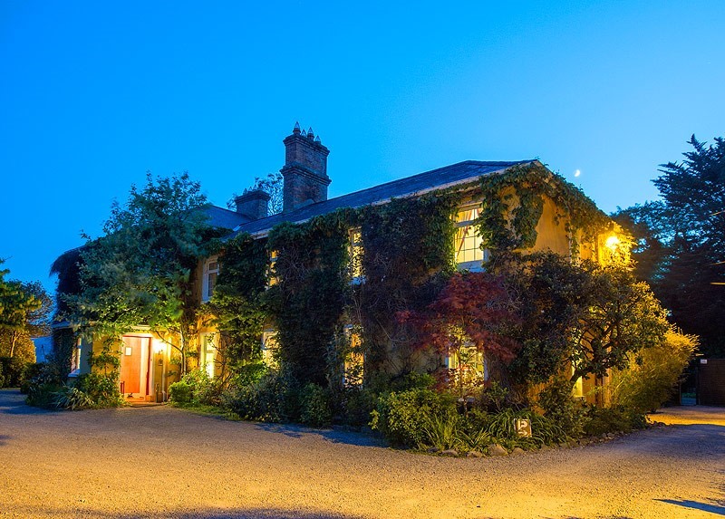 Verlicht groot huis in de schemering, omgeven door klimop en weelderig groen. Warm licht van ramen en open deur creëert een gastvrije sfeer. Schoorstenen zichtbaar op het dak. De lucht is helder en blauw, wat de gezellige ambiance versterkt. Grind oprit en goed onderhouden tuin aan de voorkant. | Echt Ierland Vakanties