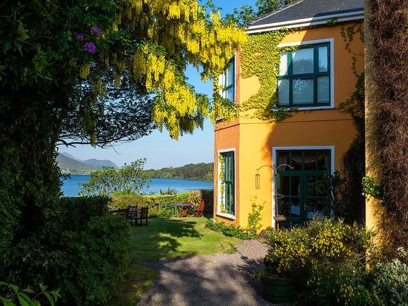 Een levendig geel huis met klimop die langs de muren klimt, staat in een weelderige tuin met uitzicht op een sereen meer. Felgele bloemen hangen aan een boom en tuinmeubilair staat op een grasveld. De achtergrond toont bergen onder een helderblauwe lucht, wat een vredig, schilderachtig uitzicht creëert. | Echt Ierland Vakanties