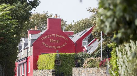 Een levendig rood gebouw, Casey's of Baltimore Hotel and Seafood Restaurant, is omgeven door weelderig groen. De ingang heeft een stenen muur en een klein zwart bord met de tekst "The Deck is Open." Omlijst door bomen ontvouwt zich dit uitnodigende tafereel onder een heldere hemel. | Echt Ierland Vakanties