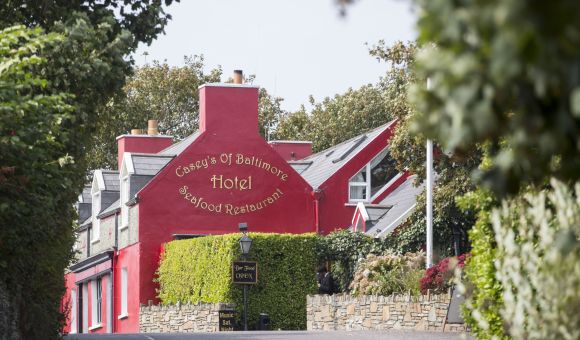 Een levendig rood gebouw, Casey's of Baltimore Hotel and Seafood Restaurant, is omgeven door weelderig groen. De ingang heeft een stenen muur en een klein zwart bord met de tekst "The Deck is Open." Omlijst door bomen ontvouwt zich dit uitnodigende tafereel onder een heldere hemel. | Echt Ierland Vakanties