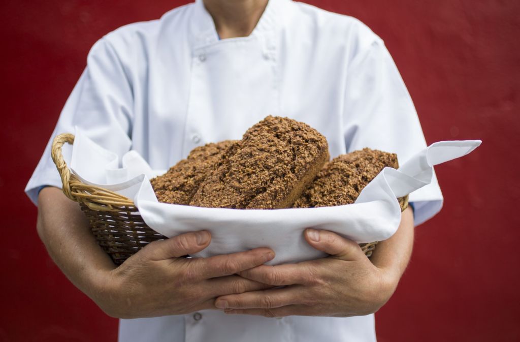 Een persoon in een witte koksjas houdt een rieten mand vast die gevuld is met verschillende broden meergranenbrood in Hotel Casey's in Baltimore. De mand, bekleed met een witte doek, steekt af tegen de dieprode achtergrond en benadrukt de versgebakken broden. | Echt Ierland Vakanties