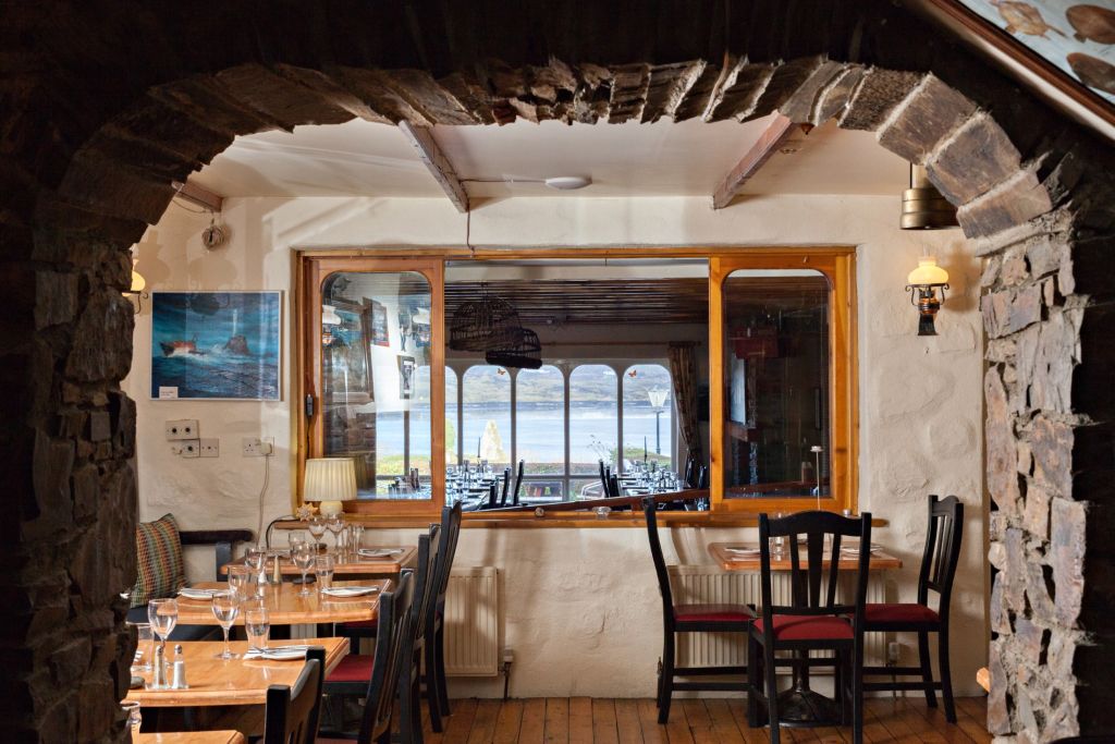 Ontdek via een stenen boog een gezellig restaurant in Hotel Casey's met houten tafels die elegant zijn gedekt met glazen en bestek. Een groot raam biedt een schilderachtig uitzicht op de zee en heuvels van Baltimore. Zachte verlichting en een rustiek decor creëren warmte, terwijl ingelijste foto's de muren sieren. | Echt Ierland Vakanties