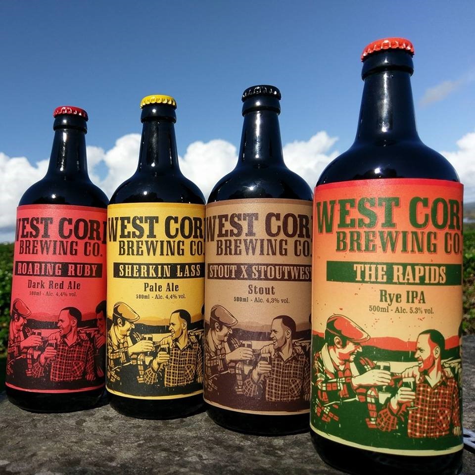 Vier flessen West Cork Brewing Co. bier staan trots buiten tegen een blauwe lucht. Met etiketten als Roaring Ruby en Sherkin Lass, toont elke fles vintage illustraties. Perfect om te genieten in Baltimore's eigen Hotel Casey's, deze brouwsels bieden aparte kleuren en ontwerpen die het oog boeien. | Echt Ierland Vakanties