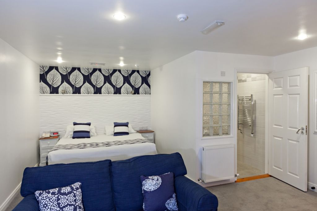 Ervaar het comfort van Hotel Casey's in Baltimore, waar een moderne slaapkamer wacht met een tweepersoonsbed versierd met blauwe en witte kussens. Een zwart-witte accentmuur met bladmotief vult de ruimte aan, terwijl een blauwe bank en kussens met patroon stijl toevoegen. Een open deur leidt naar een strakke betegelde badkamer. | Echt Ierland Vakanties