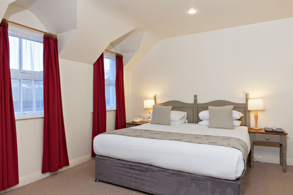 Een gezellige kamer in Hotel Casey's in Baltimore biedt een kingsize bed met wit linnen, twee grijze kussens en een beige sprei. Twee nachtkastjes met lampen flankeren het bed. Rode gordijnen sieren twee ramen aan de linkerkant en de muren zijn wit geverfd. Een telefoon en notitieblok rusten op het nachtkastje aan de rechterkant. | Echt Ierland Vakanties