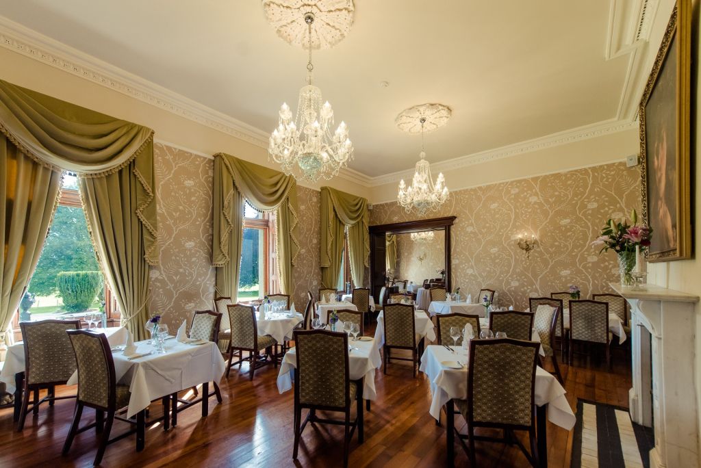 Elegante eetkamer met grote ramen met groene gordijnen, hardhouten vloeren en crèmekleurige muren. Kroonluchters hangen aan het plafond. Tafels zijn gedekt met witte kleden en formele tafelsettings. Een grote spiegel leunt tegen de verste muur en weerspiegelt het uitzicht op de tuin. | Echt Ierland Vakanties
