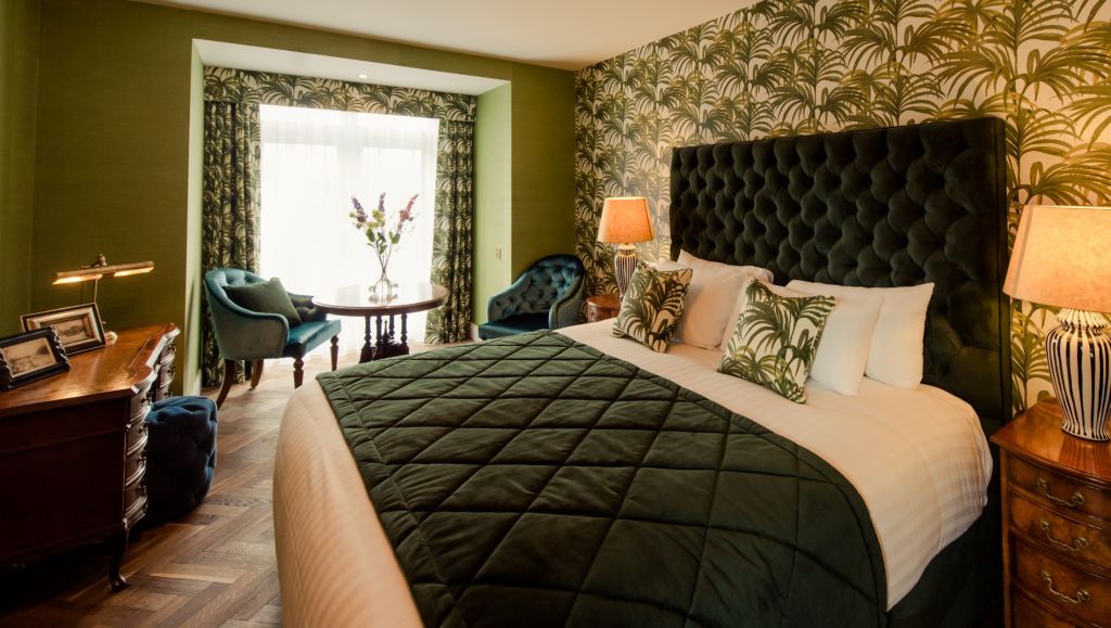 Luxe hotelkamer met een groot bed met een donkergroen dekbed en kussens met patroon. De kamer heeft groen behang met palmmotief, een houten bureau met een lamp en een zithoek bij een raam met twee stoelen en een kleine ronde tafel. Warme verlichting verlicht de ruimte. | Echt Ierland Vakanties