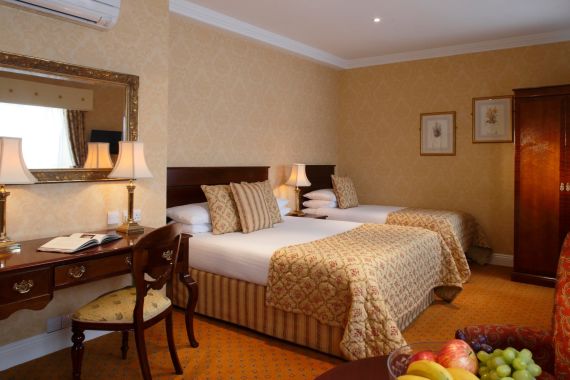 Hotelkamer met twee eenpersoonsbedden, elk met een patroonbedsprei en bijpassende kussens. Een houten bureau met een stoel en lamp staat links. Een fruitschaal siert een ronde tafel op de voorgrond. Een grote kledingkast en ingelijste kunst verfraaien de muren en bieden een ambiance die net zo elegant is als het Witte Huis zelf. | Echt Ierland Vakanties