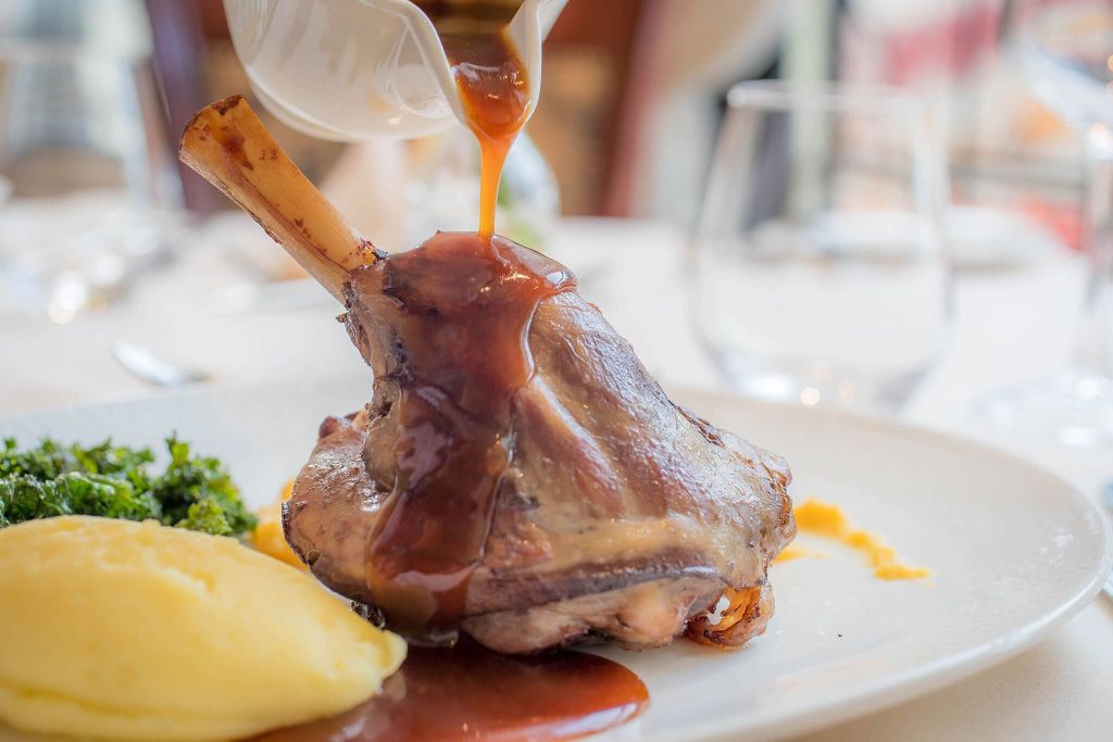 Een sappige lamsbout besprenkeld met rijke bruine jus ligt op een wit bord in het hotelrestaurant. Vergezeld van romige aardappelpuree en levendige groene groenten, staat het gerecht op een tafel met delicaat glaswerk in de wazige achtergrond, wat een elegante eetsfeer creëert. | Echt Ierland Vakanties