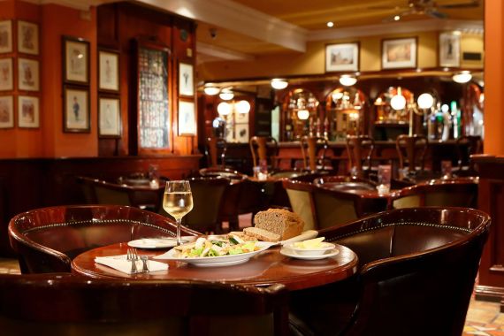 Een gezellig restaurant met gedimde verlichting en houten decor, dat doet denken aan een schilderachtig hotel. Een tafel op de voorgrond heeft een bord met gesneden brood, boter en een schaal salade met groenten en kip. Er staat een glas witte wijn naast. De achtergrond onthult meer tafels, stoelen, ingelijste kunst en een bar. | Echt Ierland Vakanties