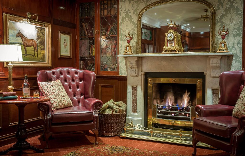 Gezellige hotelkamer met een brandende open haard, geflankeerd door sierlijke decoraties. Twee bordeauxrode leren fauteuils en een ronde tafel met een fles, glas en geruit kussen creëren een warme ambiance. Een ingelijste paardenschilderij hangt boven houten lambrisering en een mand met houtblokken staat bij de open haard. | Echt Ierland Vakanties