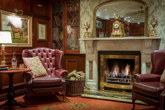 Gezellige hotelkamer met een brandende open haard, geflankeerd door sierlijke decoraties. Twee bordeauxrode leren fauteuils en een ronde tafel met een fles, glas en geruit kussen creëren een warme ambiance. Een ingelijste paardenschilderij hangt boven houten lambrisering en een mand met houtblokken staat bij de open haard. | Echt Ierland Vakanties