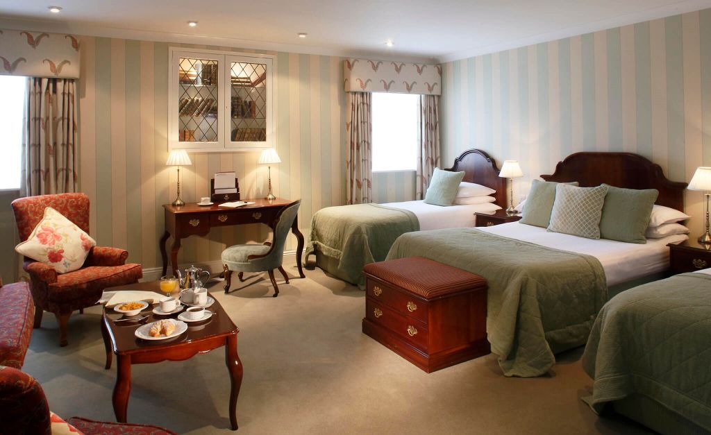 In een hotelkamer die doet denken aan het Witte Huis, staan twee eenpersoonsbedden met groen beddengoed en voldoende kussens. Een houten bureau rust tegen gestreept behang, terwijl een kleine tafel met ontbijtartikelen wordt geflankeerd door gestoffeerde stoelen. Lichte gordijnen voor de ramen laten natuurlijk licht de ruimte sieren. | Echt Ierland Vakanties