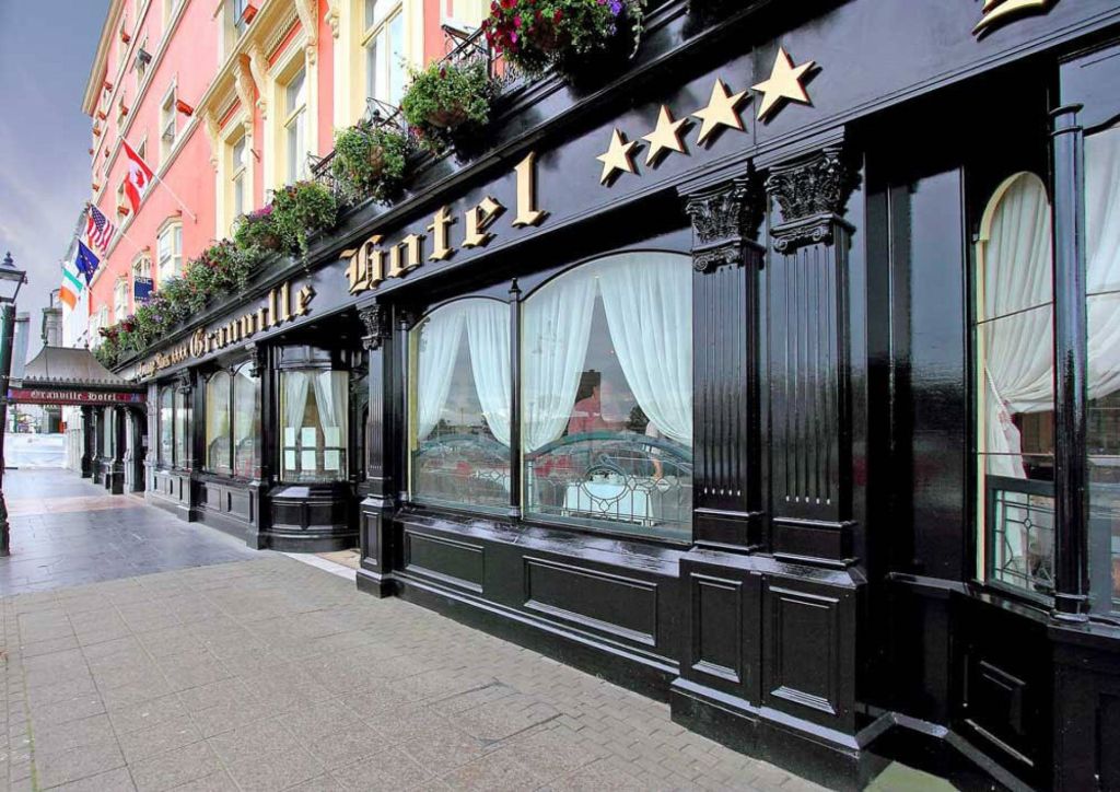 Elegant hotel exterieur met een zwarte façade en grote ramen met witte gordijnen. Gouden letters spellen "Hotel" onder vier sterren. Er hangen vlaggen boven, die doen denken aan het Witte Huis, terwijl een roze bovenmuur contrast toevoegt. Een geplaveid trottoir ligt op de voorgrond. | Echt Ierland Vakanties