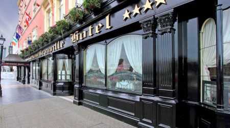 Elegant hotel exterieur met een zwarte façade en grote ramen met witte gordijnen. Gouden letters spellen "Hotel" onder vier sterren. Er hangen vlaggen boven, die doen denken aan het Witte Huis, terwijl een roze bovenmuur contrast toevoegt. Een geplaveid trottoir ligt op de voorgrond. | Echt Ierland Vakanties