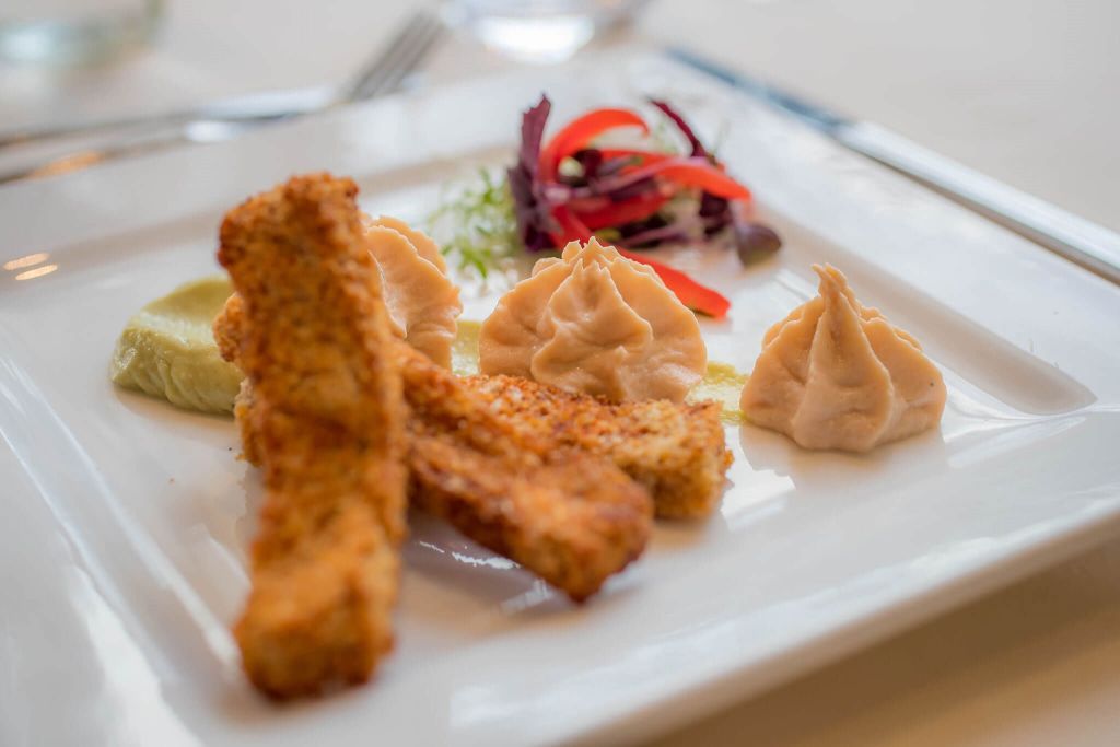 Een gastronomisch gerecht op een vierkant wit bord uit onze hotelkeuken bestaat uit drie gepaneerde visstaafjes, verschillende scheppen romige beige saus, een smeersel van groene puree en een kleine garnering van bladsalade met rode peperreepjes. De achtergrond is zacht vervaagd, waardoor het opgediende eten wordt benadrukt. | Echt Ierland Vakanties