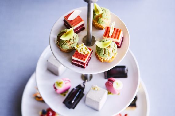 Een dessertkraam met drie lagen bevat een assortiment aan gebak en taarten op een tafel. De bovenste laag bevat kleurrijke taarten, de middelste heeft gelaagde taarten en de onderste toont verschillende zoetigheden. Op de achtergrond staan twee borden met extra gebakjes die wazig zijn gemaakt. | Echt Ierland Vakanties