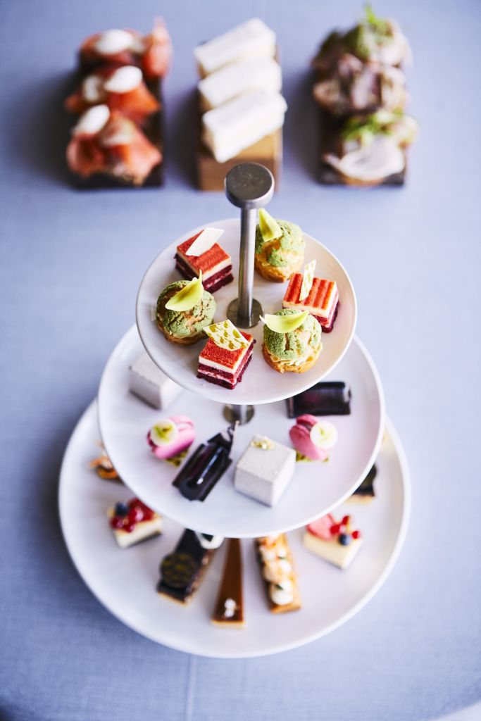 Een dessertkraam met drie lagen bevat een assortiment aan gebak en taarten op een tafel. De bovenste laag bevat kleurrijke taarten, de middelste heeft gelaagde taarten en de onderste toont verschillende zoetigheden. Op de achtergrond staan twee borden met extra gebakjes die wazig zijn gemaakt. | Echt Ierland Vakanties