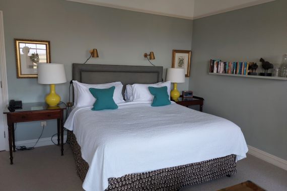 Een netjes opgemaakt bed met een witte sprei en twee blauwgroene kussens staat in het midden tegen een lichtblauwgroene muur. Twee gele lampen met witte kappen staan op houten nachtkastjes. Daarboven zijn koperen wandlampen gemonteerd. Een plank met boeken en decoratieve items staat rechts. Ingelijste kunst hangt aan de muur. | Echt Ierland Vakanties