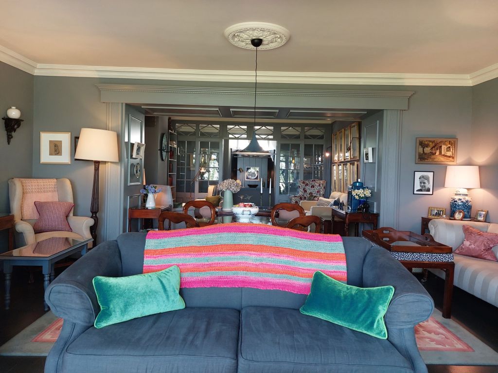 Een gezellige woonkamer met een pluche grijze bank met een kleurrijke gestreepte deken en blauwgroene kussens. De kamer is versierd met meubels in vintagestijl, waaronder fauteuils, een staande lamp en ingelijste kunstwerken. Een spiegel weerspiegelt een goed gedekte eettafel en zachte verlichting creëert een warme sfeer. | Echt Ierland Vakanties