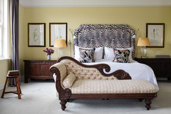 Elegante slaapkamer met een groot bed met een zebra-patroon hoofdbord en bloemenkussens. Een klassieke chaise longue met gecapitonneerde bekleding staat op de voorgrond. Ingelijste botanische prints hangen aan de lichtgele muren. Twee lampen met beige kappen sieren donkere houten nachtkastjes. | Echt Ierland Vakanties
