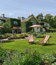 Een weelderige tuin met twee gestreepte ligstoelen op een verzorgd gazon op de voorgrond. Een schilderachtig huisje, bedekt met klimop, staat op de achtergrond met grote ramen en verschillende tafels met parasols. Bloemperken met kleurrijke bloemen omzomen het gazon onder een helderblauwe lucht. | Echt Ierland Vakanties
